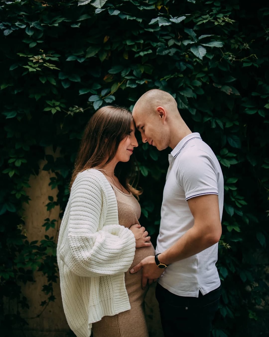 Expecting couple standing close together in front of deep green foliage